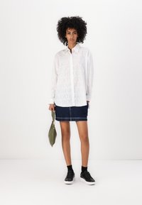White, long-sleeve button-up shirt with repeated "HUGO" logo in light print, paired with a dark denim skirt and black sneakers. Holding a green bag.