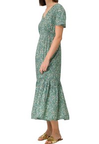 Femme portant une robe midi fleurie bleu canard à manches courtes et ourlet froncé, associée à des sandales plates ouvertes dorées.