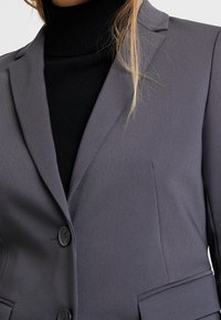 Blazer gris sur mesure avec deux boutons, revers à encoches et tissu texturé. Porté par-dessus un col roulé noir, il présente une coupe ajustée et des poches.