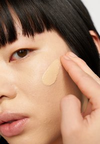 Fond de teint beige appliqué sur la peau avec une texture lisse, estompé avec les doigts. Gros plan sur un visage avec des cheveux courts noirs et une couleur de lèvres naturelle.