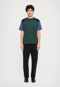 Uomo in piedi contro uno sfondo bianco, che indossa una maglietta a maniche corte verde, viola e nera, pantaloni neri e scarpe nere.