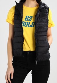 Czarna pikowana kamizelka puchowa z zamkiem z przodu i kapturem, nałożona na żółtą koszulkę z krótkim rękawem z niebieskim nadrukiem, w zestawie z czarnymi spodniami.