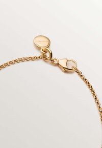 Pulsera de cadena de oro con un cierre tipo langosta y una etiqueta redonda con bordes acanalados. Textura suave con diseño entrelazado.