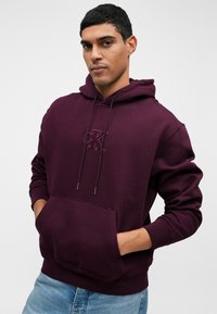 Hanorac cu glugă de culoare vișinie, fabricat dintr-un amestec de bumbac, dotat cu buzunar frontal, șnur de ajustare și logo embossat pe piept. Material moale cu manșete ribbate.