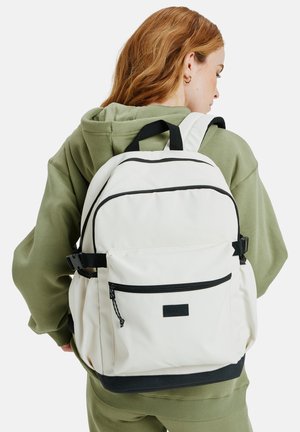 Biały plecak z tkaniny z czarnymi suwakami i akcentami, regulowanymi szelkami i przednią kieszenią, noszony na oliwkowym zielonym hoodie.
