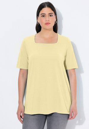 Frau mit langen dunklen Haaren trägt ein blassgelbes Kurzarm-Top mit quadratischem Ausschnitt, kombiniert mit grauer Hose, stehend vor weißem Hintergrund.