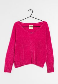 Pull tricoté rose avec col en V et manches longues et larges. Présente des poignets et un ourlet côtelés. Suspendu à un accroche noir sur un fond blanc.