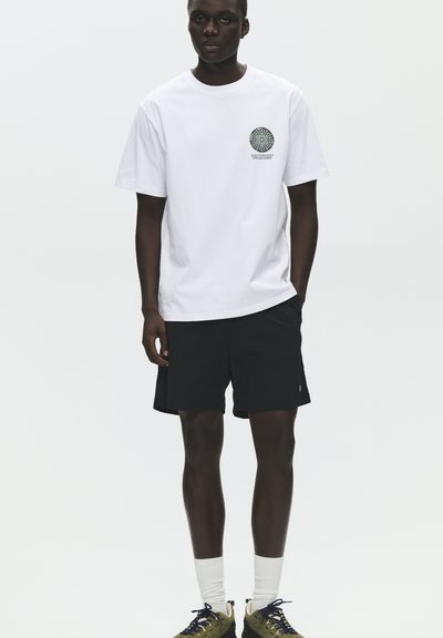 Άνδρας στέκεται φορώντας λευκό T-shirt με σχέδιο, μαύρο σορτς, λευκές κάλτσες και μπεζ αθλητικά παπούτσια με μαύρα κορδόνια σε απλό φόντο.