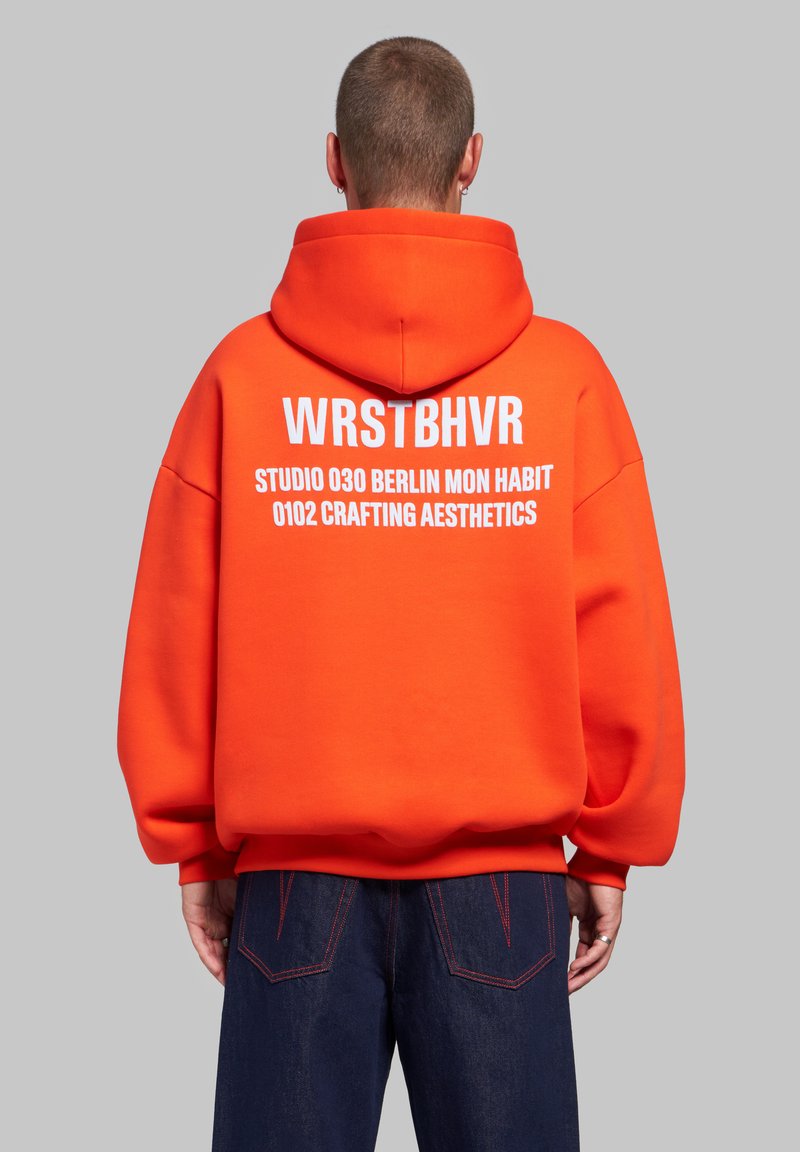 Personne portant un sweat à capuche orange vif avec un texte blanc "WRSTBHVR STUDIO 030 BERLIN MON HABIT 0102 CRAFTING AESTHETICS" dans le dos, associé à un jean foncé.