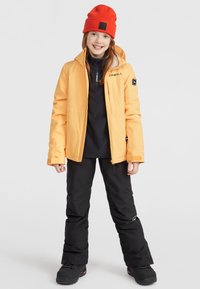 Orange vandtæt jakke med lynlås, sort termisk skjorte og sorte snesokker. En rød hue og sorte støvler fuldender outfitet.