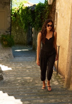 Femme en tenue noire et lunettes de soleil marchant sur des marches en pierre le long d’un vieux bâtiment ensoleillé avec de la verdure en arrière-plan.