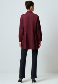 Woman with hair bun wearing burgundy long-sleeve tunic, navy pants, black heels, standing facing a plain light background.
