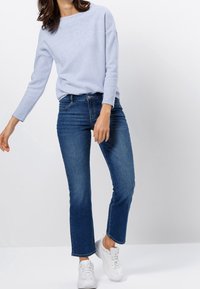 Femme portant un pull à manches longues bleu clair, un jean bleu taille mi-haute et des baskets blanches, se tenant devant un fond clair.