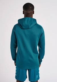 Felpa con cappuccio color teal a maniche lunghe, texture morbida e fit rilassato, abbinata a pantaloni corti teal coordinati con logo sulla gamba.