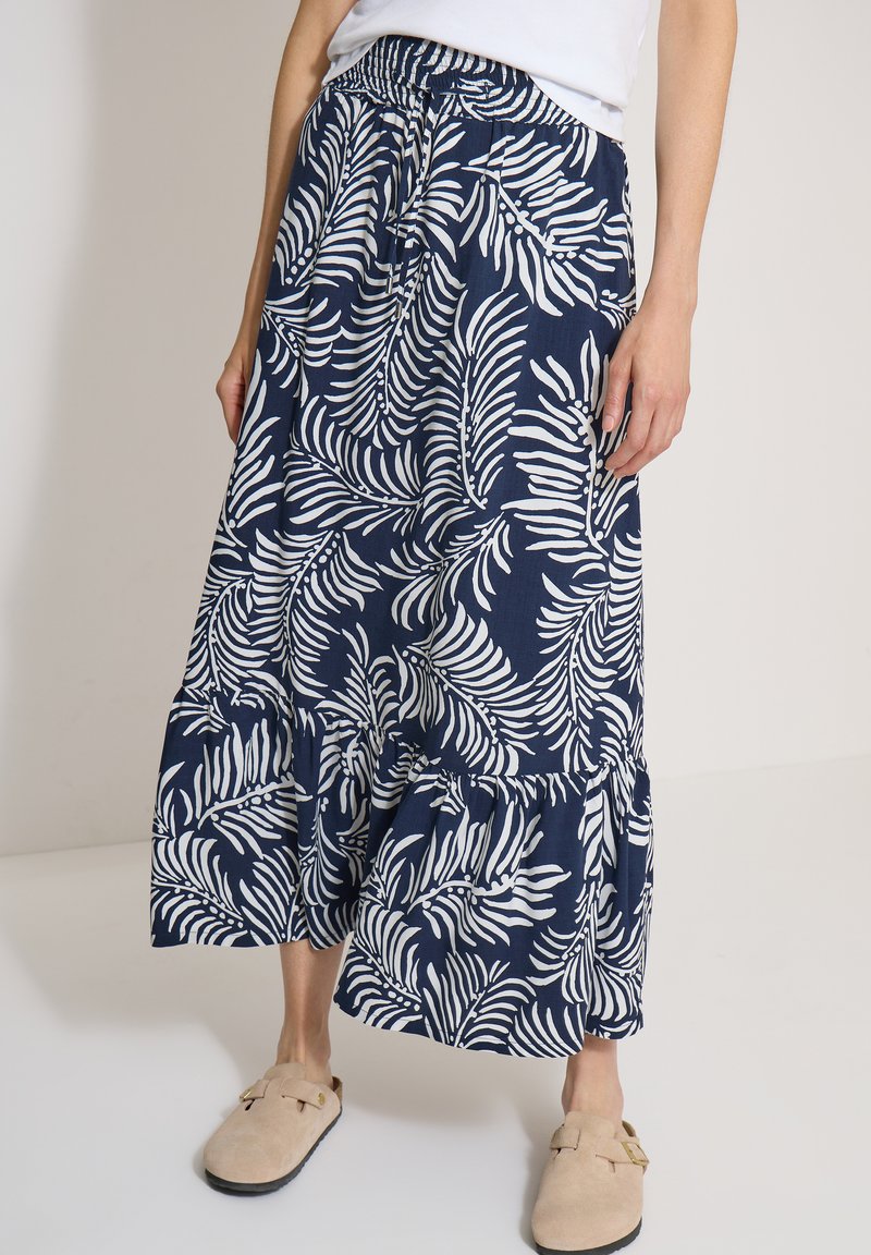 Femme portant une jupe midi bleu marine et blanc à motif feuilles avec des chaussures beiges slip-on, debout devant un fond uni.