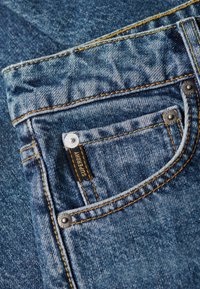 Niebieskie dżinsy z przodu z kieszenią, ozdobione pomarańczowymi szwami, metalowymi elementami i czarną metką z wyraźnym napisem "Superdry".