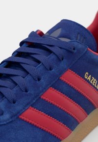 Nærbillede af blå ruskinds sneakers med røde striber, blå snørebånd, gummisål og guld "GAZELLE" branding på siden.