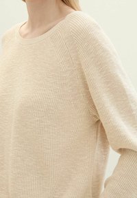 Beżowy sweter robiony na drutach z szerokim dekoltem, rękawami raglanowymi i teksturowanym wzorem. Materiał wygląda na miękki i lekki.