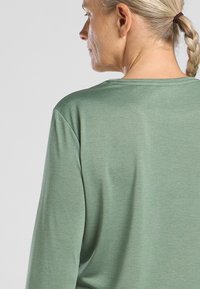 Langärmliges, Rundhalsoberteil in Hellgrün, aus weichem, strukturiertem Stoff mit minimalen Nähten und lockerem Schnitt. Die Rückansicht hebt die schlichte Gestaltung hervor.