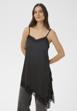 Frau trägt ein schwarzes Seiden-Camisole mit Spitzenbesatz und graue Jeans, steht mit den Händen hinter dem Rücken vor einem schlichten hellen Hintergrund.