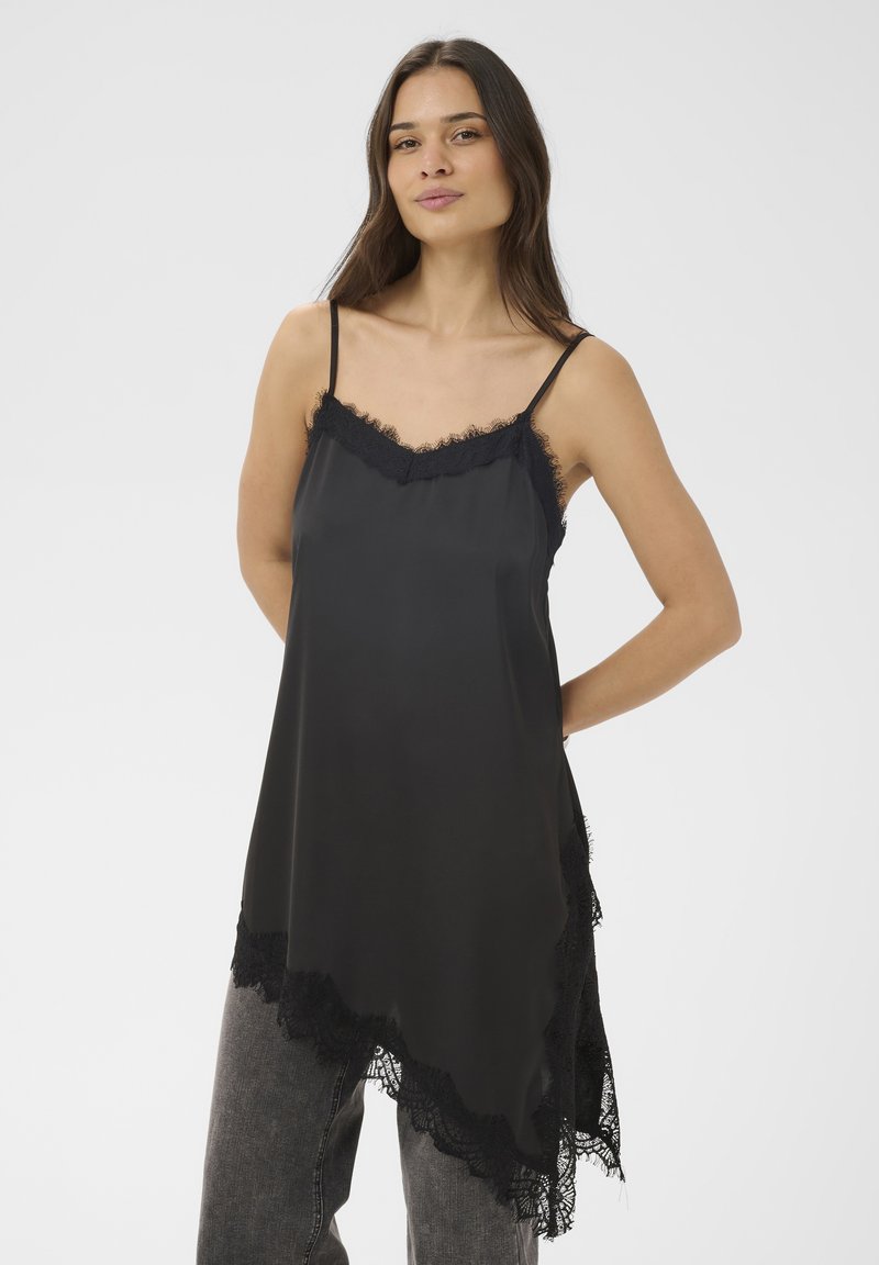 Femme portant un caraco en soie noire avec bordure en dentelle et jean gris, debout, les mains derrière le dos, sur un fond clair uni.