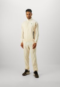 Light beige track suit with a zip-up jacket and elastic cuffs, featuring side pockets with black zippers and a small logo. Matched with dark shoes.
