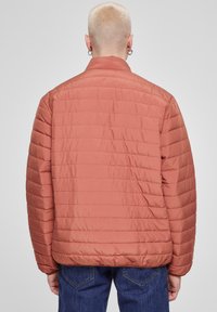 Puffjakke i dempet korallfarge, med horisontale sømmer, lange ermer og ståkrage. Stoffet ser glatt og lett ut.