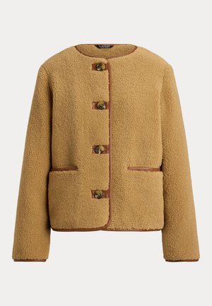 Femme portant une veste polaire couleur beige sur une tenue en denim foncé, tenant un sac à main en cuir marron, debout sur un sol en bois clair contre un mur uni.