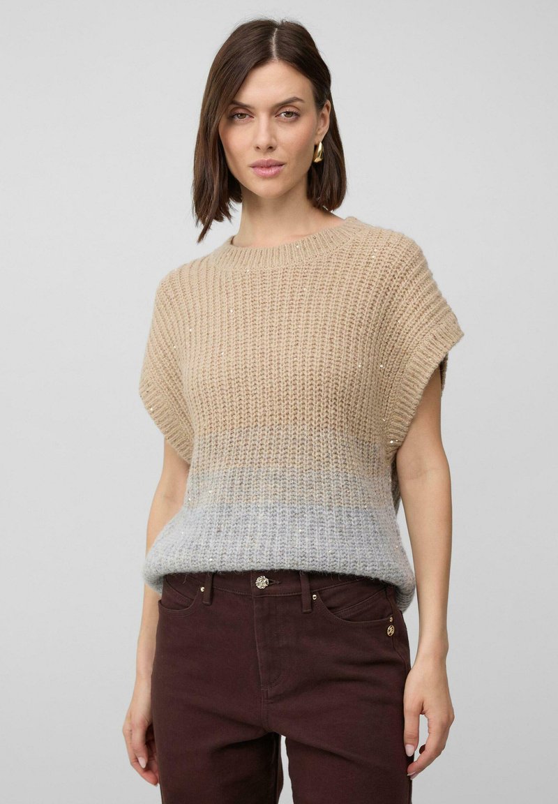 Vrouw met kort bruin haar, draagt gouden oorringen, een beige-grijze gebreide zonder mouwen trui en donkerbruine broek tegen een effen achtergrond.