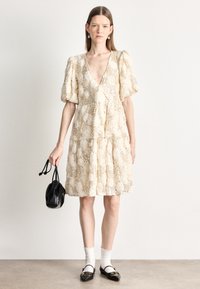 Μπεζ φόρεμα με λουλούδια, βαθύ V-λαιμόκοψη, φουσκωτά μανίκια και φούστα με βολάν. Φορεμένο με μαύρη μικρή τσάντα, άσπρες κάλτσες με βολάν και μαύρα φλατ παπούτσια.