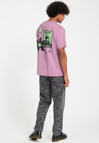 Roze katoenen t-shirt met een grafisch ontwerp op de achterkant met een groene glimlachende gezicht, sterren en abstracte patronen. Gepaard met zwarte broeken met een patroon.