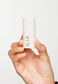 ILIA stickverpakking in een matte crèmekleur, cilindervorm met een lichtgrijs logo. Vastgehouden in een hand die een gladde huidtextuur toont.