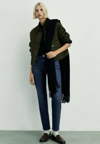 Olivgrön jacka med brun krage, svart scarf, mörkblå jeans och bruna loafers. Outfiten har en minimalistisk design och texturerad tyg.