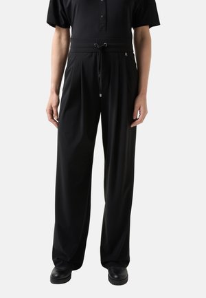 Osoba nosząca czarne, szerokie spodnie z sznurkiem w pasie, w połączeniu z czarnymi butami i czarną bluzką z krótkim rękawem zapinaną na guziki.