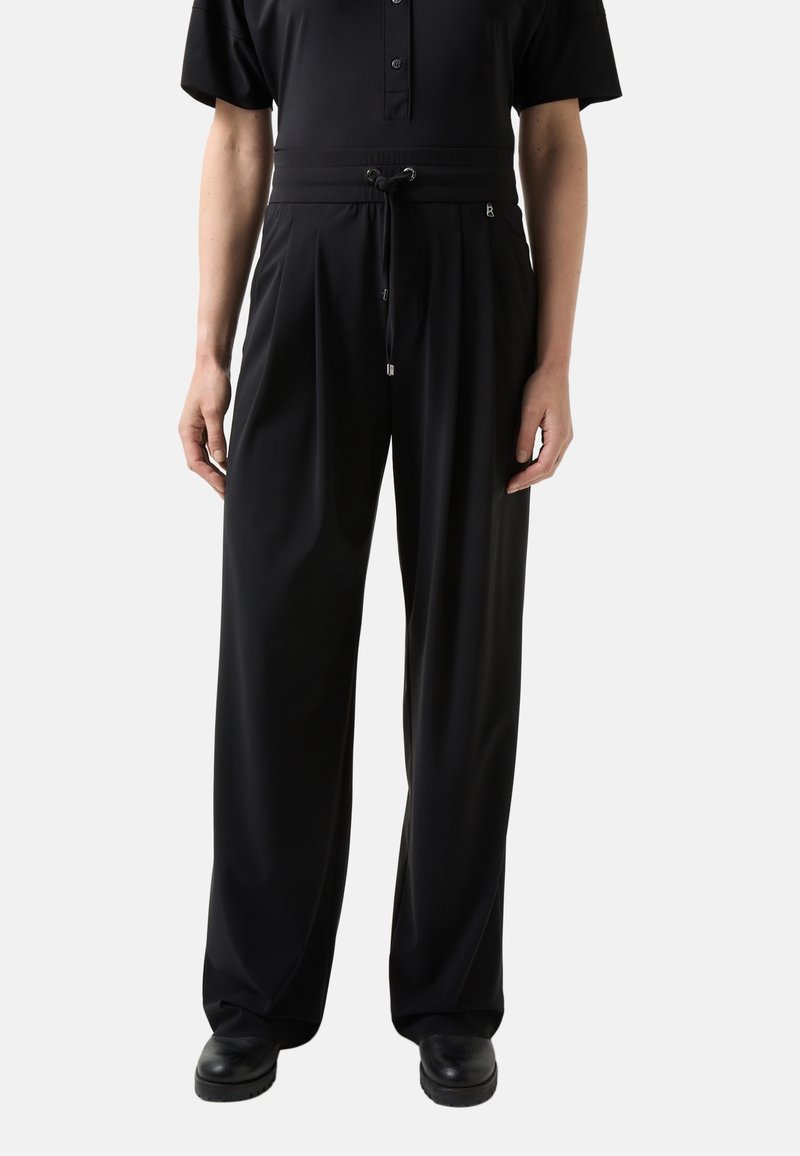 Personne portant un pantalon noir à jambes larges avec une taille à cordon, assorti à des chaussures noires et un haut noir à manches courtes avec des boutons.