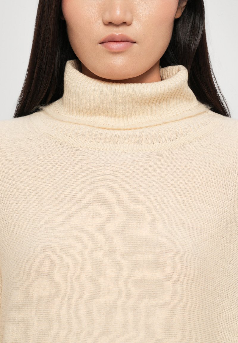 Pull col roulé crème avec texture côtelée et plis au niveau du col. Matière douce, design ajusté, style minimaliste.