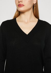 Sweater preto em V com mangas longas, feito de material de malha texturizada. Apresenta um decote em V profundo e um corte descontraído. Detalhes subtis nas bordas.