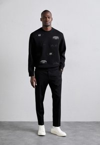 Črna majica s teksturiranimi obliži, podrobnostmi logotipa in okroglo izrezom. Ujemajoča se z črnimi hlačami in belimi supergami, stoječa pred nevtralno kuliso.