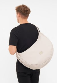 Borsa a tracolla in morbido tessuto beige con forma arrotondata, tracolla regolabile e un piccolo marchio. Indossata sulla spalla di una persona.
