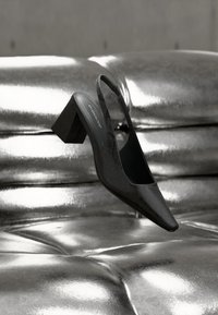 Μαύρο υφασμάτινο γόνατο με ψηλό τακούνι και ιμάντα στον αστράγαλο, τοποθετημένο πάνω σε γυαλιστερή ασημένια επιφάνεια από δερμάτινο ταπετσαριστό ύφασμα.