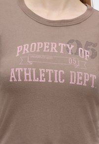 Близък план на кафява риза с розов текст, гласящ "PROPERTY OF ATHLETIC DEPT" и по-малък текст "UNIVERSITY DIST. 05" на гърдите.