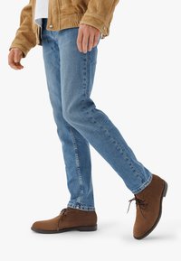 Bottines en daim marron avec un bout arrondi et des détails à lacets, associées à un jean ajusté bleu clair et une veste beige.