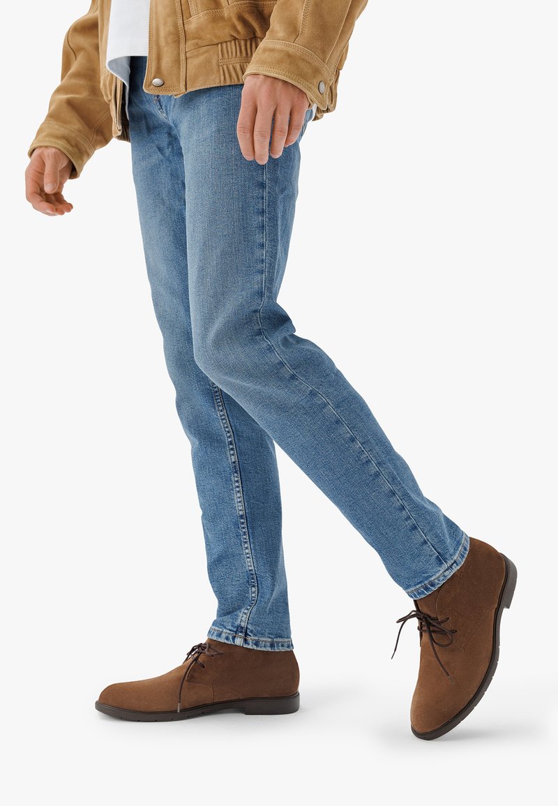 Bottines en daim marron avec un bout arrondi et des détails à lacets, associées à un jean ajusté bleu clair et une veste beige.