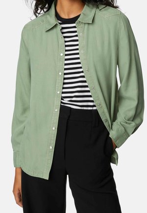 Camisa de botones verde claro, hecha de una tela suave, con mangas largas, llevada sobre una camiseta a rayas blancas y negras, combinada con pantalones negros.
