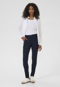 White long-sleeve shirt over a white tank top, paired with navy fitted pants and white sneakers with tan accents. Smooth fabric, casual look.