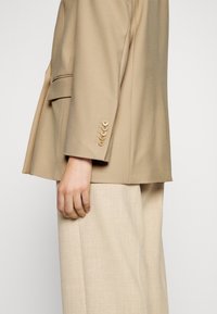 Blazer beige con un ajuste entallado, que presenta un bolsillo único y seis botones en los puños, combinado con pantalones de pierna ancha en beige claro.