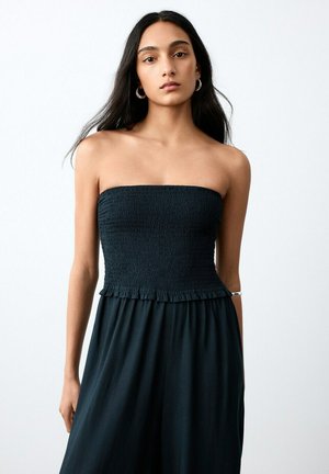 Giovane donna con capelli lunghi e scuri, che indossa un vestito nero senza bretelle con corpetto arricciato, orecchini a cerchio argentati e braccialetto d'argento sul braccio scoperto.