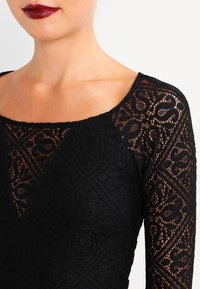 Zwarte kanten bodystocking met lange mouwen en een vierkante halslijn. Bevat ingewikkelde patronen en een gestructureerde afwerking. De halslijn heeft een subtiele V-vorm.