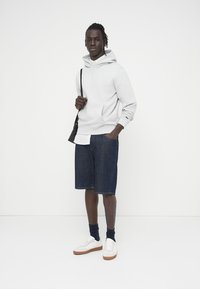 Homme debout portant un sweat à capuche blanc, un short en jean foncé, des chaussettes bleu marine, des baskets blanches, et portant un sac à bandoulière noir sur un fond blanc uni.