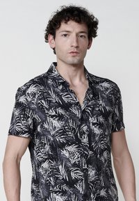 Camisa de botón de manga corta en negro con un estampado de hojas en gris y blanco, que presenta un cuello tipo campamento y un sutil logo en el pecho.